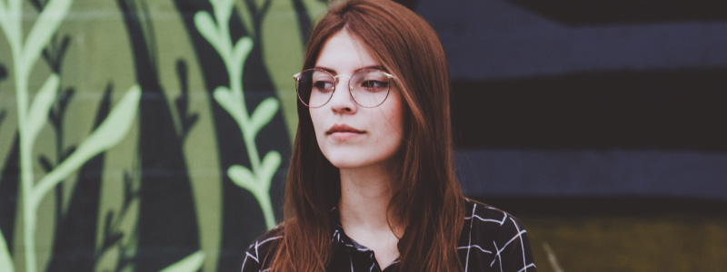 young woman with auburn hair and large glasses standing in front of a mural and looking off to the side
