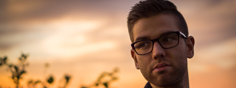 brunette man wearing glasses looking off to side, sunset in the back