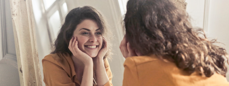 brunette woman wearing yellow shirt, looking in mirror and smiling