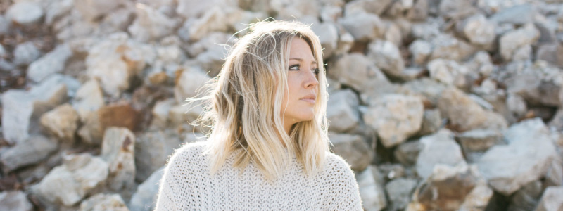 Blonde woman wearing a white sweater in front of a wall of rocks considering her swollen gums
