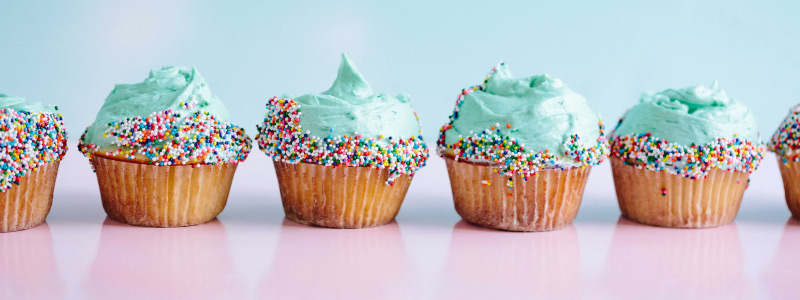 Row of vanilla cupcakes with blue frosting and colorful sprinkles on a pink counter against a blue wall