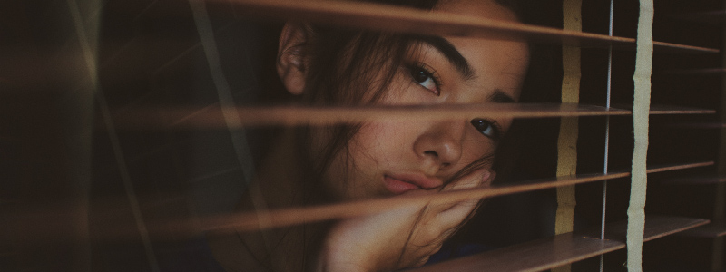 Brunette woman rests her cheek on her hand due to jaw pain while looking sadly through window blinds