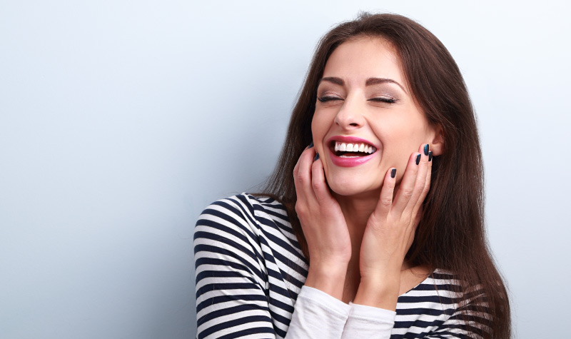 Brunette woman with plenty of saliva wears a black and white striped shirt while smiling and touching her cheeks