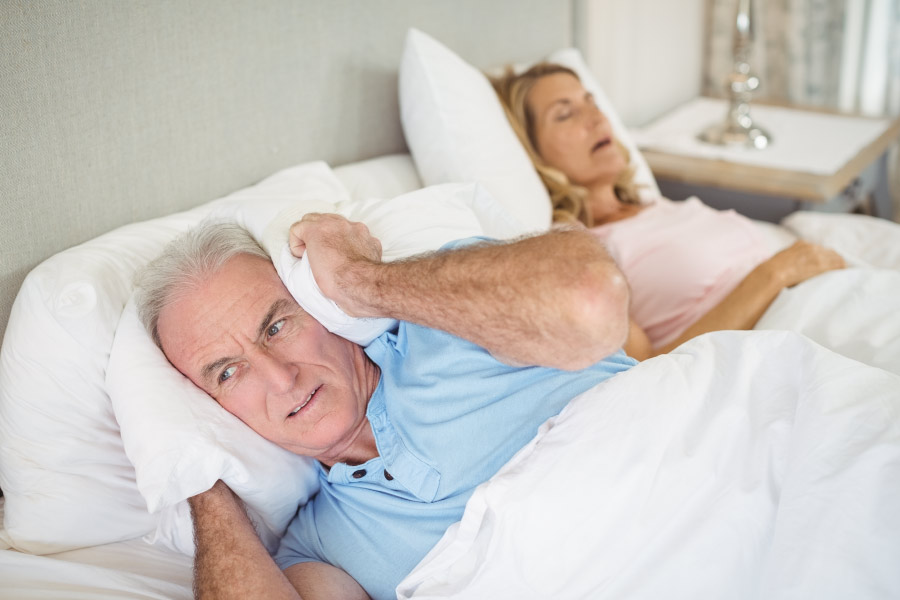 Man holding a pillow over his ears while his wife sleeps & snores next to him due to sleep apnea